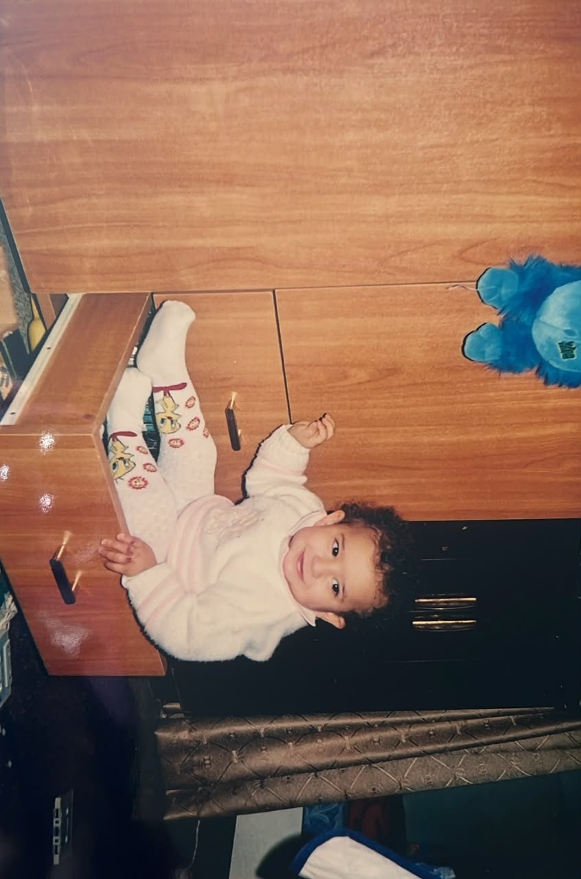 An old printed photograph of Fatima as a baby in a white jumper, sitting on an open drawer and smiling at the camera.