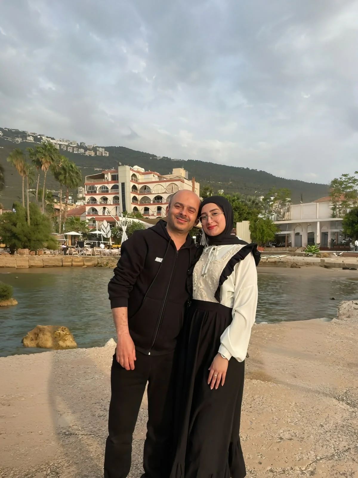 Fatima leaning against a man in a black hoodie by the water, hills and palm trees behind them.