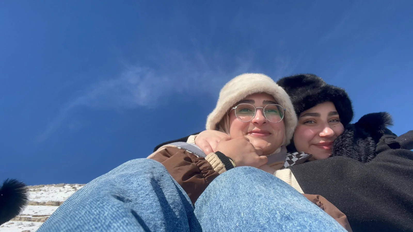 Two bundled-up faces cheek to cheek under a deep blue winter sky, snow on the ground behind.