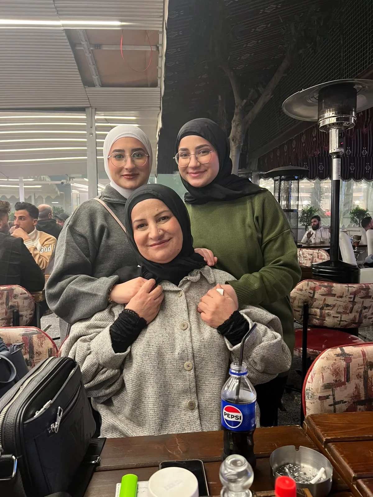 Fatima and another young woman standing behind a seated woman, arms around her, at a café table.