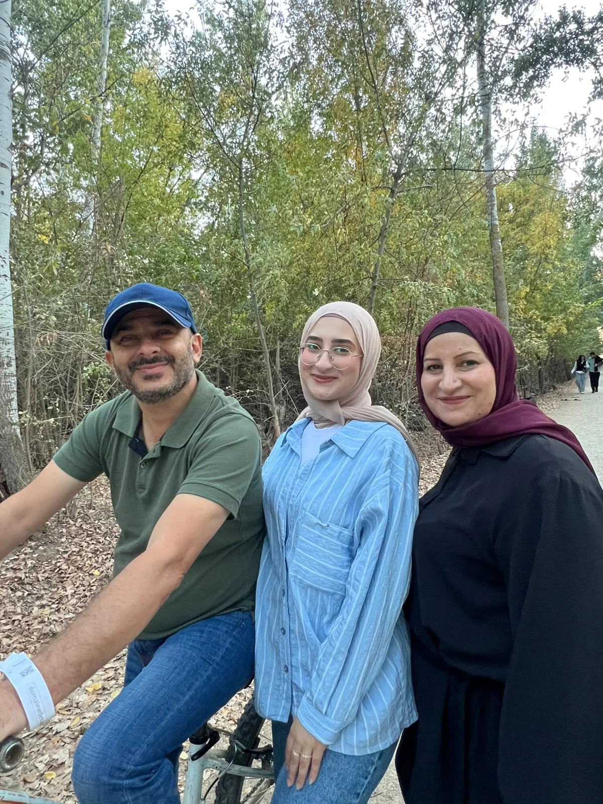Fatima on a tree-lined path, standing between a man on a bicycle and a woman in a burgundy scarf.