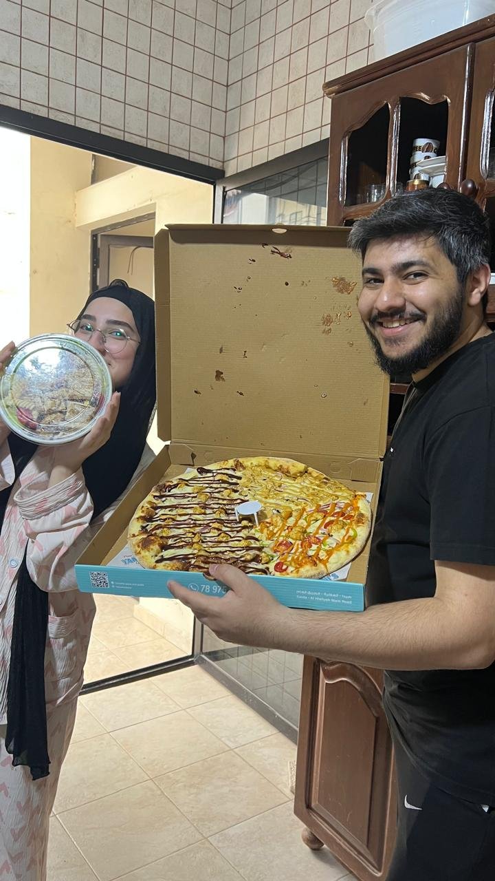 In the kitchen: an open pizza box held up for the camera while Fatima hides half her face behind a container of food.